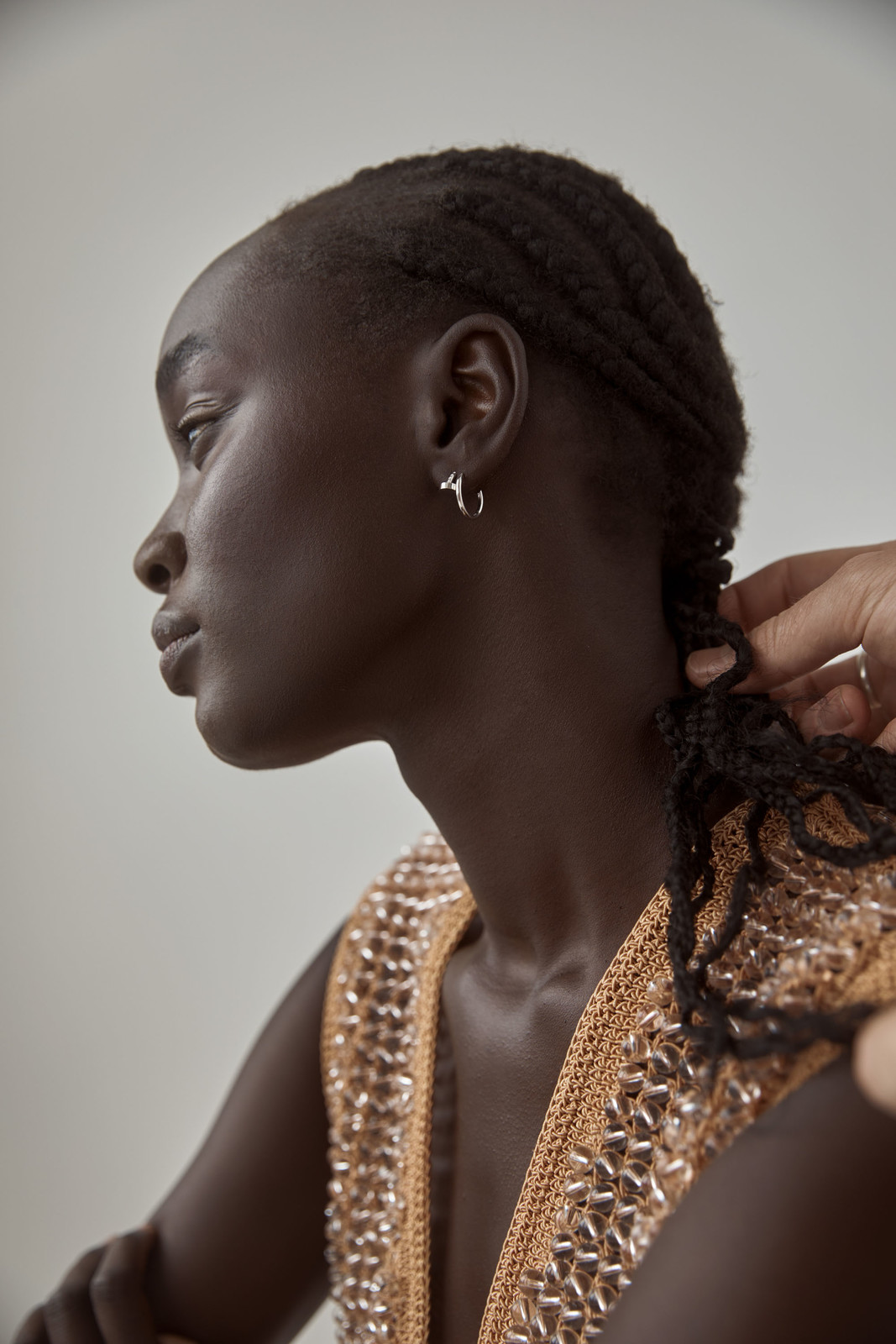 Profile shot of a dark-skinned woman with braided hair, wearing a gold top, having her hair adjusted by an unseen person, indoors.