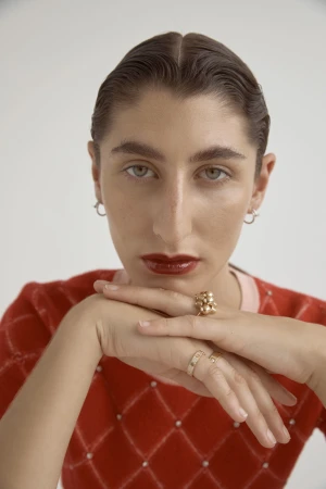 Close-up portrait of a model with dark slicked-back hair wearing a red argyle top, holding hands under chin showcasing rings.