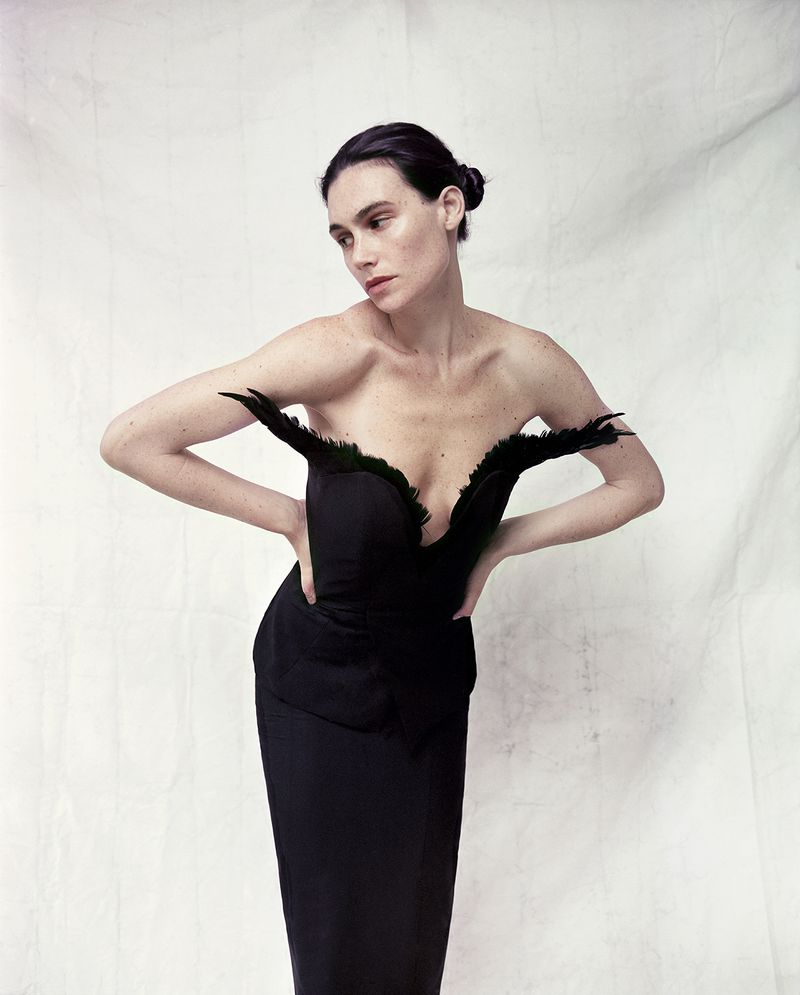 A woman in a black strapless dress with feather detailing, posing against a textured white backdrop. Fashion and portrait photography.