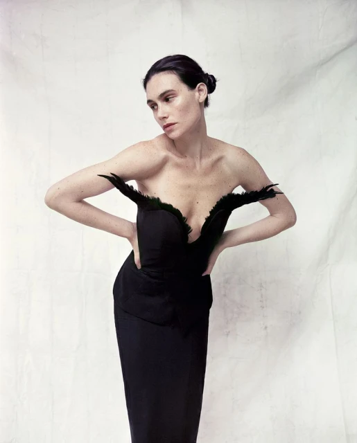 A woman in a black strapless dress with feather detailing, posing against a textured white backdrop. Fashion and portrait photography.