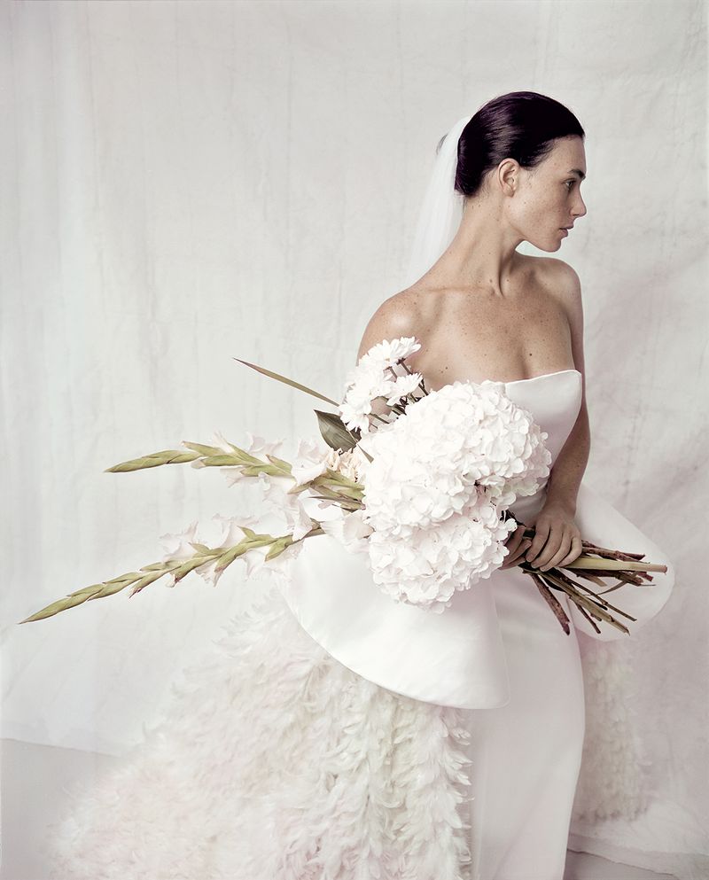 Elegant woman in white wedding dress holds a bouquet against a neutral background, with soft lighting creating a sophisticated look.