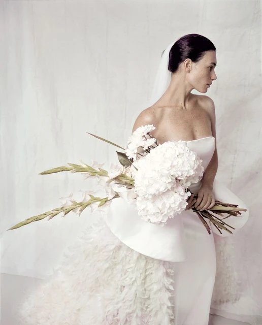 Elegant woman in white wedding dress holds a bouquet against a neutral background, with soft lighting creating a sophisticated look.