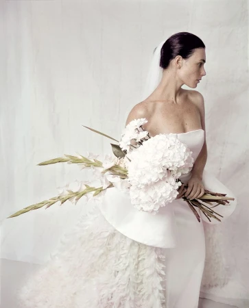 Elegant woman in white wedding dress holds a bouquet against a neutral background, with soft lighting creating a sophisticated look.