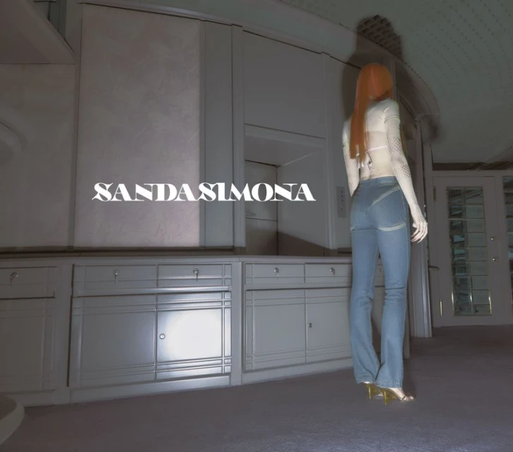 Woman in a light top and jeans stands in a room with kitchen cabinets, bathed in soft light. A blend of fashion and interior.