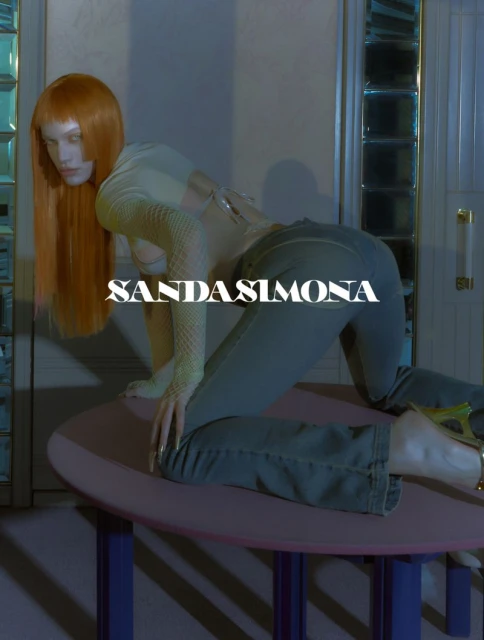 Woman with an orange wig poses on a table wearing jeans and a sheer top in a fashion style portrait against a neutral background.