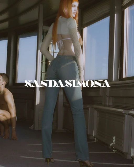 Fashion shot of a woman with orange hair wearing jeans and a cropped top, posed in front of windows, with a man in the background.