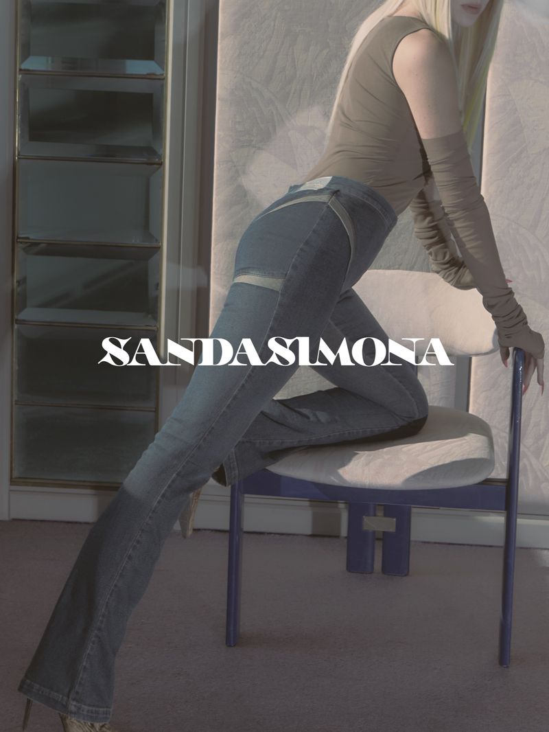 Fashion shot of a woman posing in jeans and long gloves, seated on a chair in an indoor studio with soft, diffused lighting.