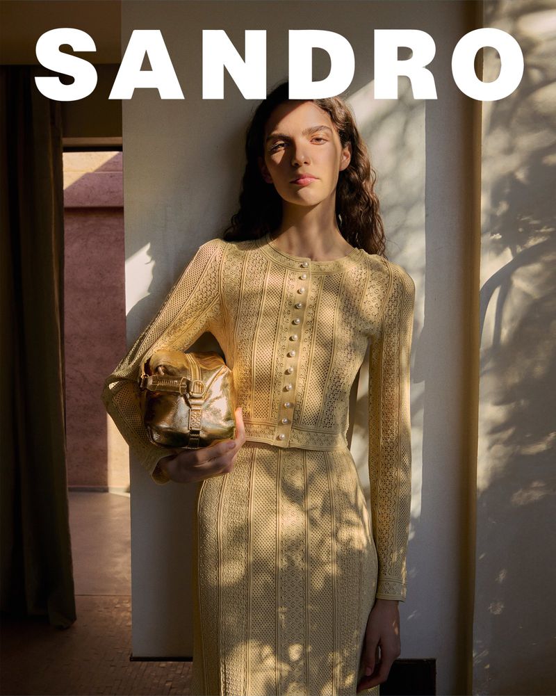 A young adult woman poses indoors in a yellow knit outfit with a handbag against a neutral background in a fashion setting.