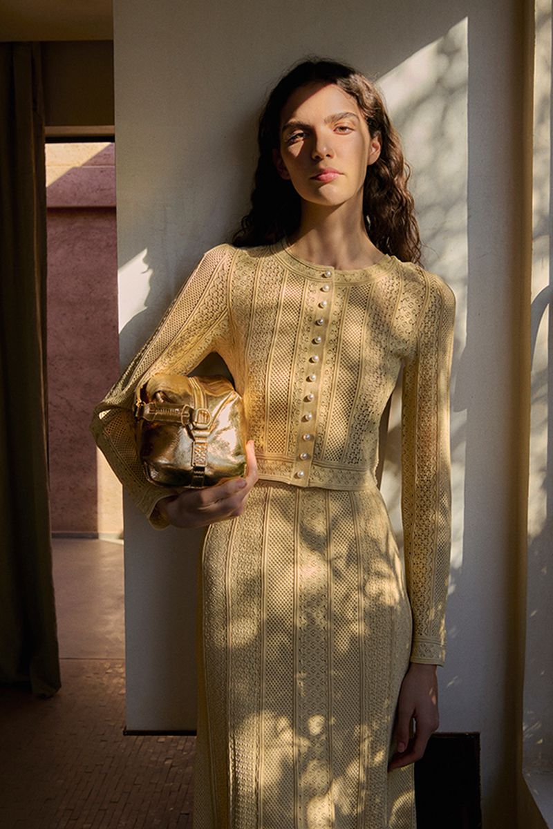 Model wearing a textured yellow knit cardigan and skirt holding a gold metallic handbag indoors
