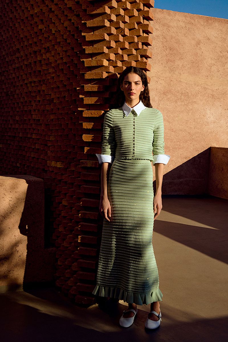 A woman stands in front of a textured wall, wearing a knit dress and Mary Jane shoes in a sunlit outdoor setting for a fashion shot.