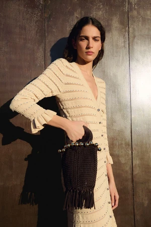 Brunette woman poses in front of a textured wall, wearing a cream-colored dress and holding a dark brown purse with fringe.