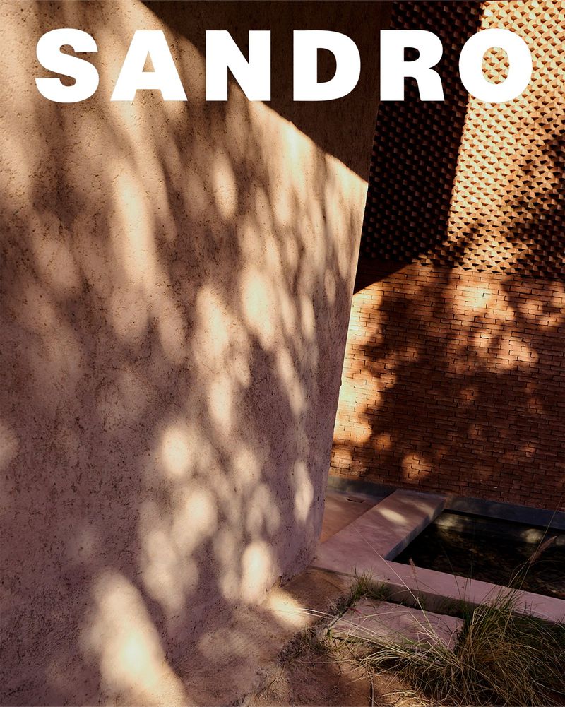 Architectural outdoor shot featuring a textured wall with dappled sunlight, a glimpse of a brick building, and a stone-lined water feature.