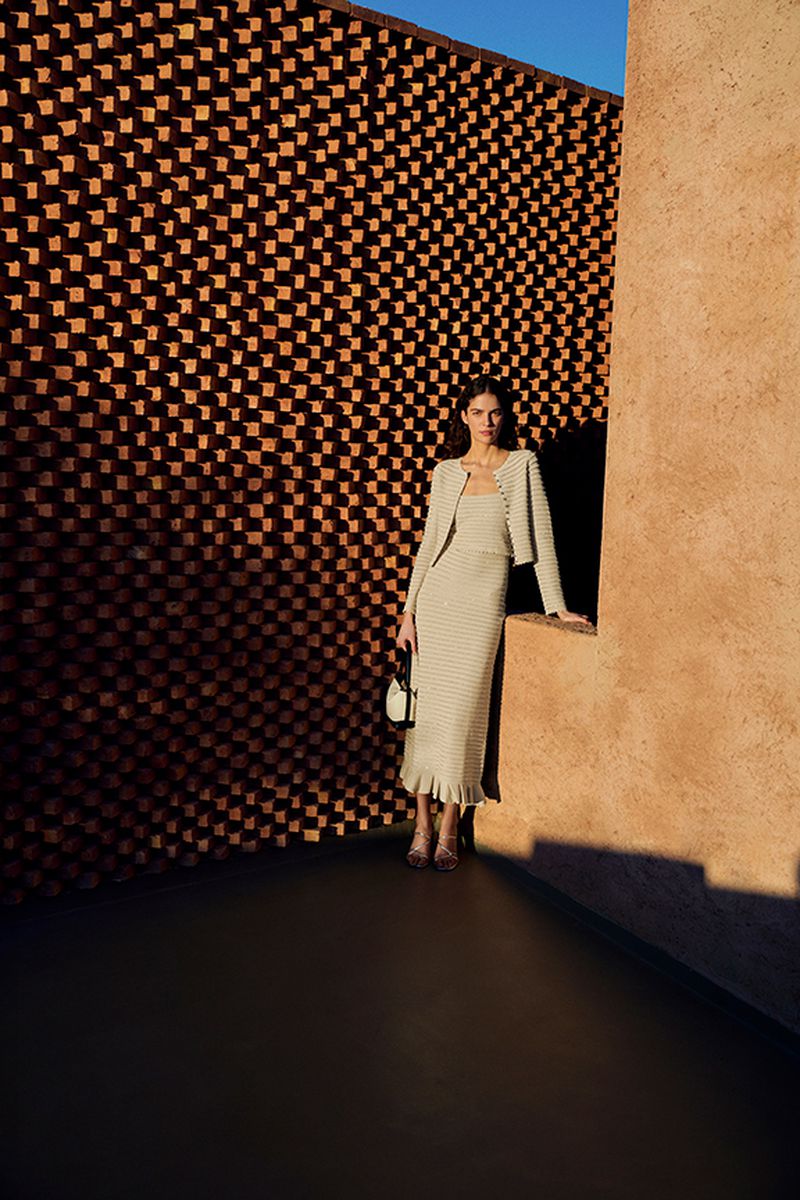 Woman in knitwear poses outdoors in neutral colors. She wears a dress, jacket, and heels and holds a small handbag in soft sunlight.