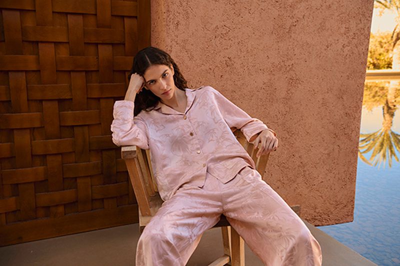 A woman in pink pajamas sits on a wooden chair outdoors against an adobe wall, with a blurred reflection of palm trees in the background.