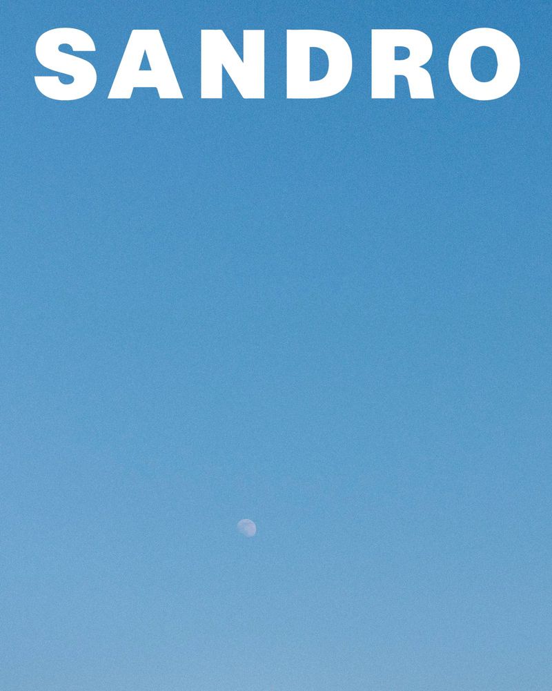 Clear blue sky with large white brand text at the top and a small crescent moon visible below for Sandro S/S 25