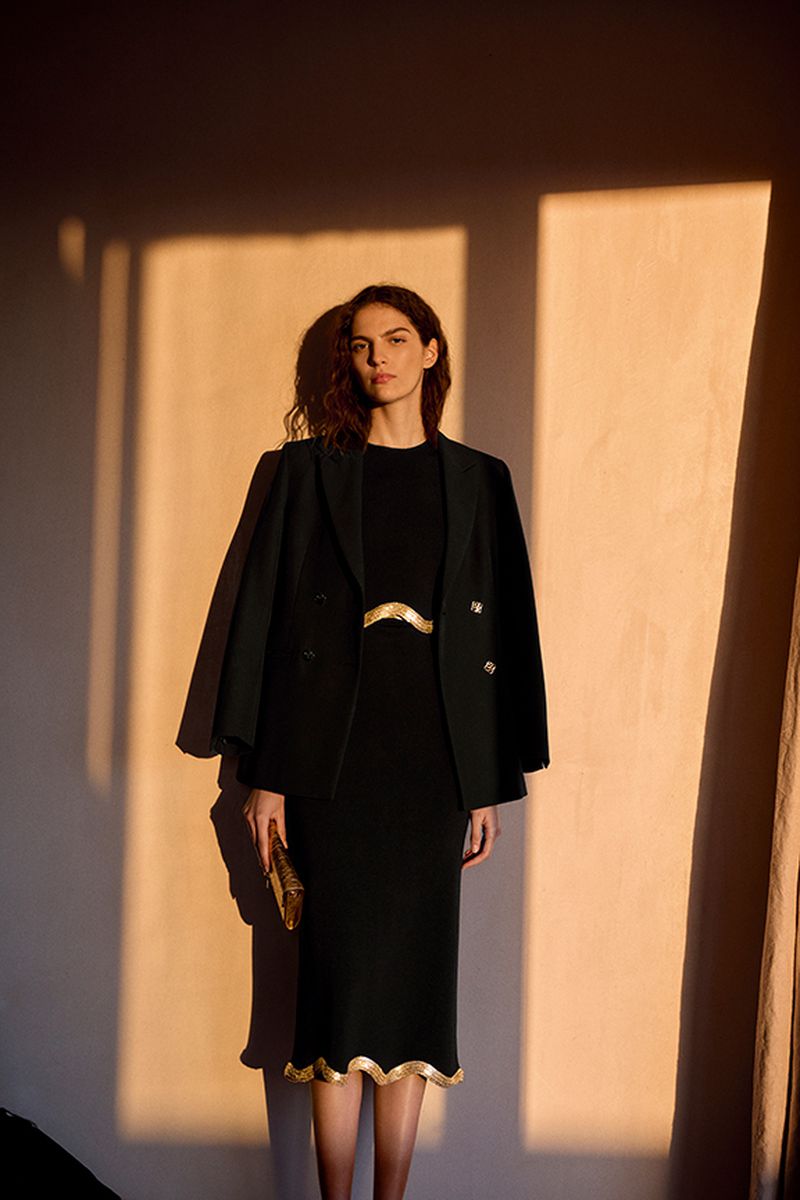 Woman in a black dress and blazer stands in front of a wall with window shadows, holding a gold clutch; a stylish and professional look.