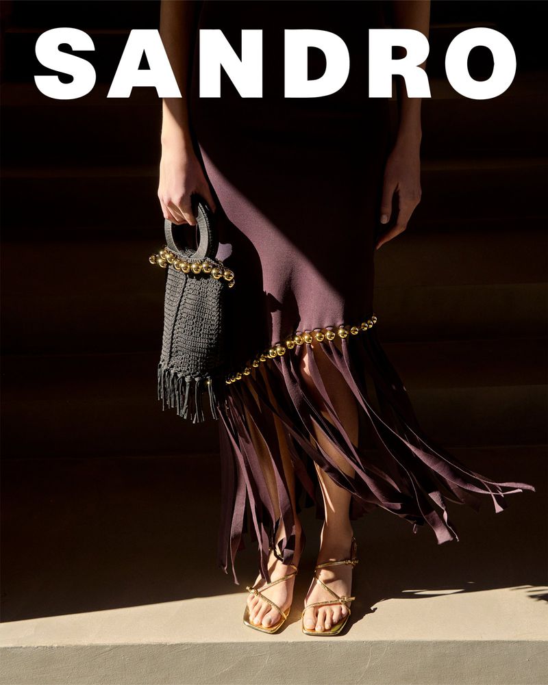 Fashion editorial shot of a woman wearing a plum colored dress, gold sandals, and a black handbag, with the brand name SANDRO visible.