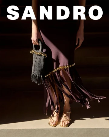 Fashion editorial shot of a woman wearing a plum colored dress, gold sandals, and a black handbag, with the brand name SANDRO visible.