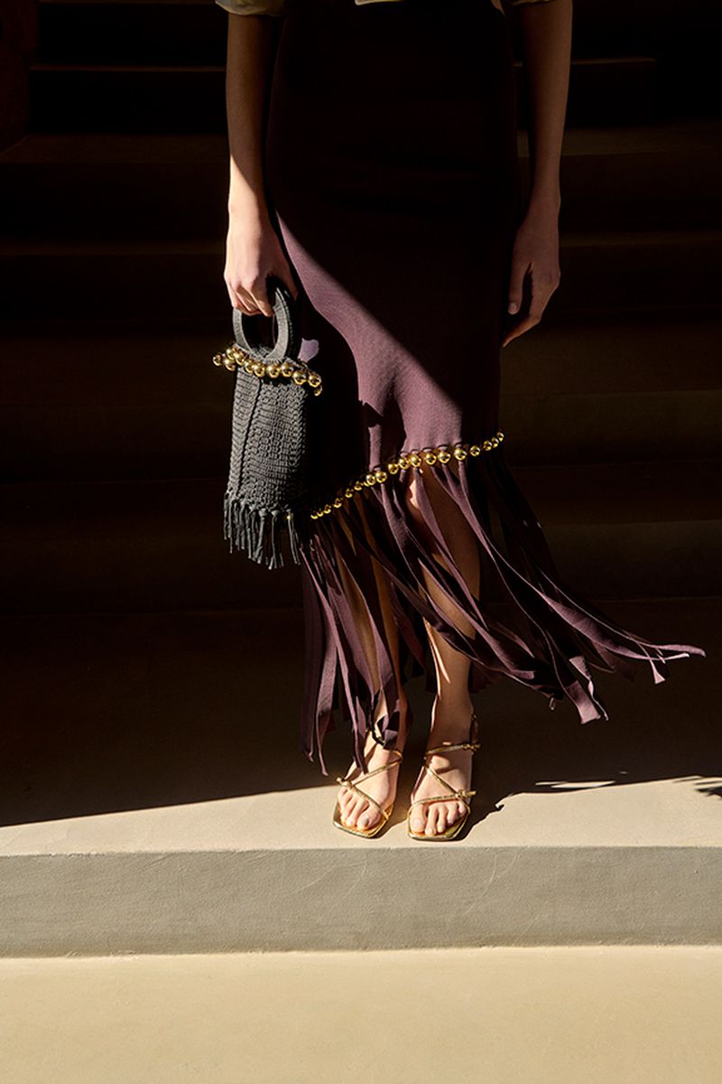 Woman in a brown skirt stands on stairs holding a woven purse with fringe and gold sandals in a naturally lit indoor setting.