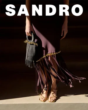 Model wearing a dark skirt with a fringed hem and gold hardware, holding a textured black handbag and strappy sandals.