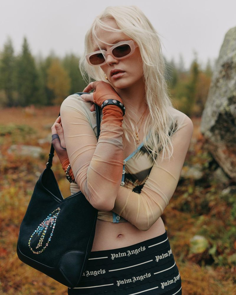 A slender woman with blonde hair and sunglasses poses outdoors, showcasing a black purse, long-sleeved top, and Palm Angels branded bottoms.