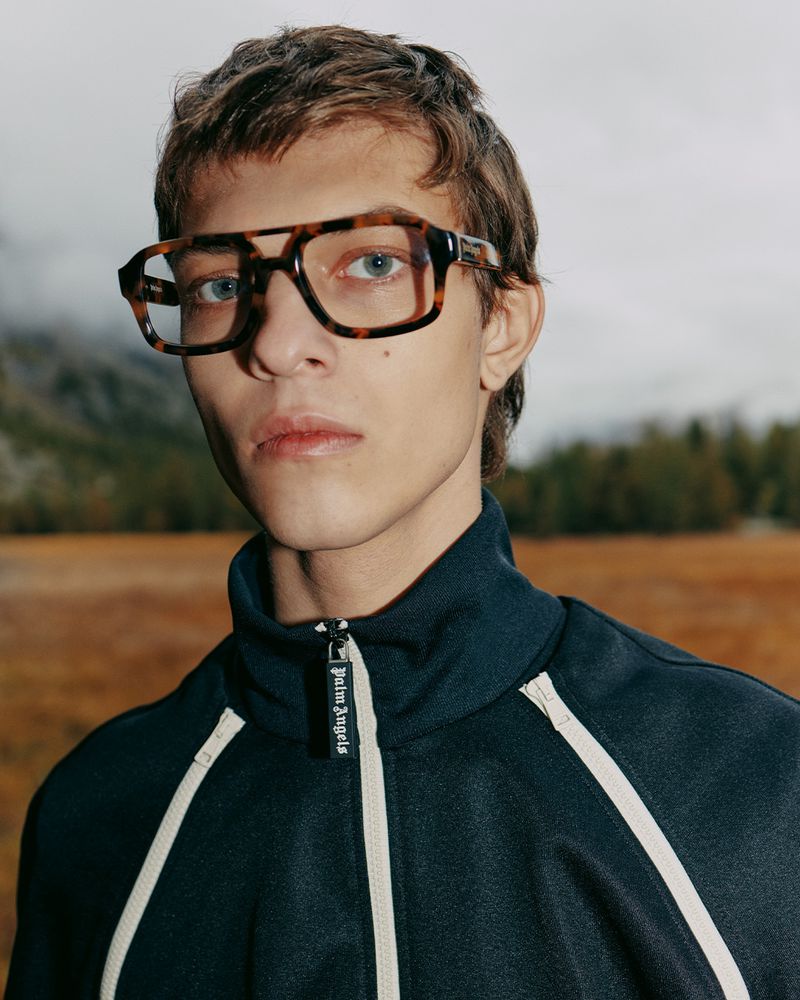 Portrait of a young adult male wearing dark track jacket and tortoise shell glasses against an outdoor, blurred natural backdrop.