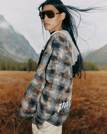 A person with long black hair wears sunglasses and a plaid jacket against a background of mountains and a field.