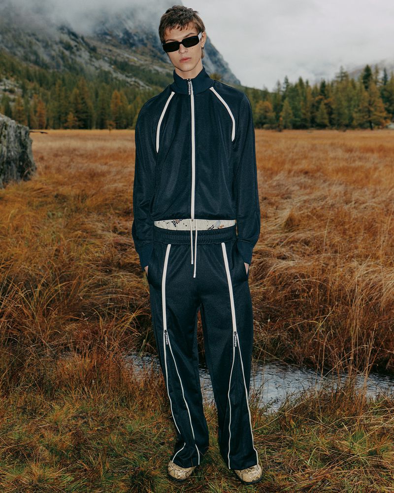 Male fashion model in a blue tracksuit poses in a field of brown grass with mountains in the background, showcasing outdoor athletic style.
