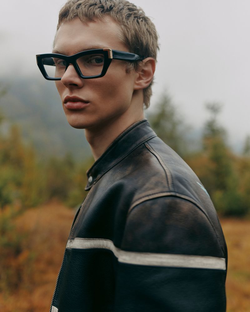 Close-up of a male fashion model wearing black-framed eyeglasses and a black leather jacket in a natural outdoor setting.