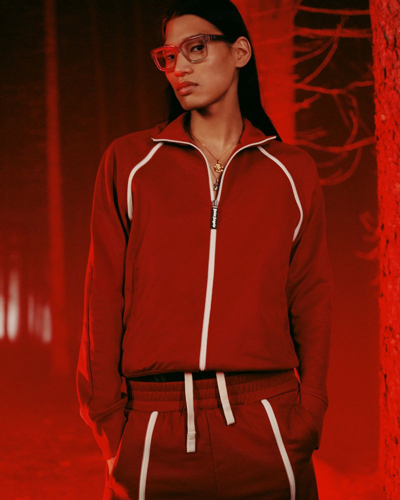 A person with glasses and a red tracksuit stands in front of trees in a reddish-lit background, creating a striking fashion-forward portrait.