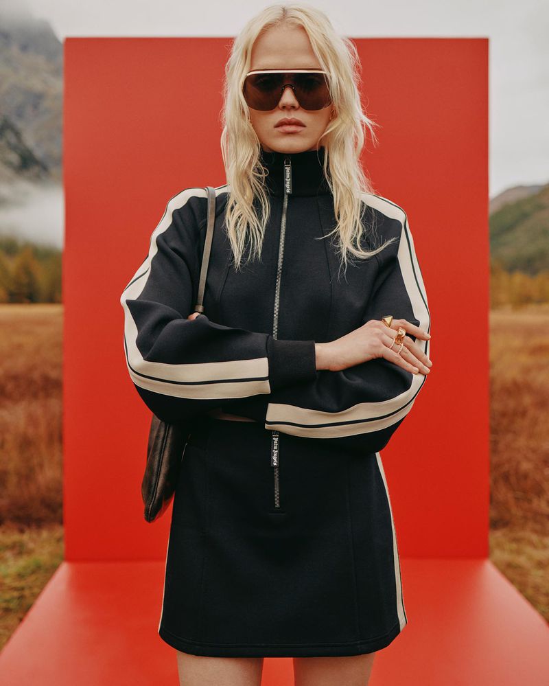 Fashion photo of a blonde woman wearing sunglasses and a black mini dress with a red backdrop and mountain landscape. Outdoors shot.