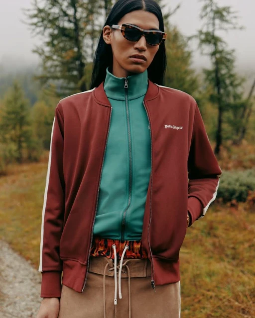 Woman in red tracksuit jacket and sunglasses poses in nature with blurred green trees in the background on an overcast day.