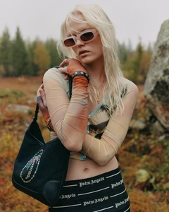 Blonde model in sunglasses wearing sheer long sleeve top holding a black shoulder bag outdoors in woodland landscape