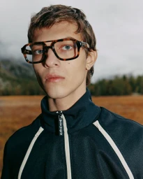 Portrait of a man wearing tortoiseshell glasses and a dark tracksuit jacket against an outdoor landscape background