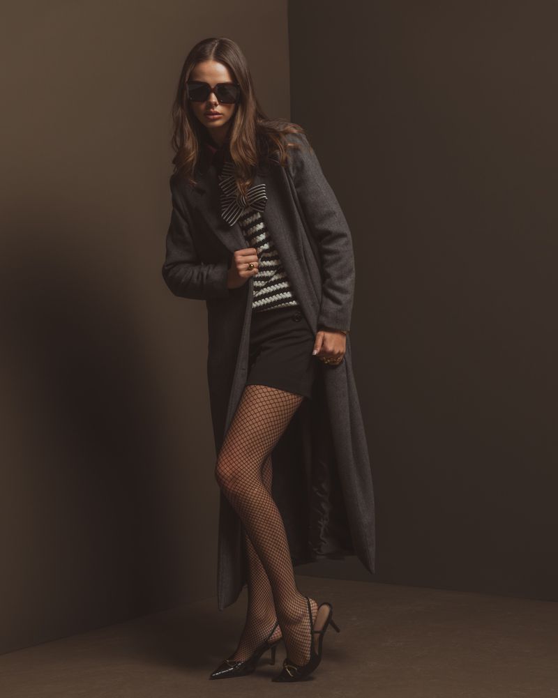A woman poses in a fashion shot wearing a gray coat, striped top, black skirt, patterned tights, and black high heels in a studio.