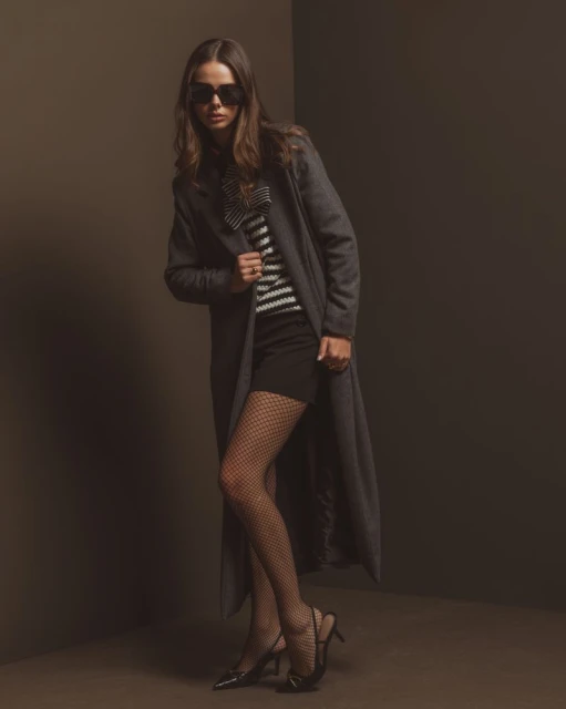 A woman poses in a fashion shot wearing a gray coat, striped top, black skirt, patterned tights, and black high heels in a studio.