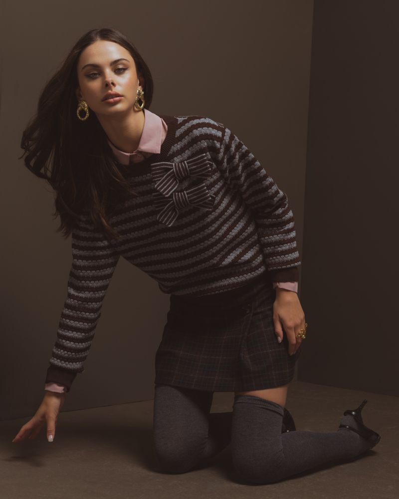 Model kneeling on a dark studio floor wearing a striped knit sweater with bow accents, pink shirt, dark skirt, and grey stockings.