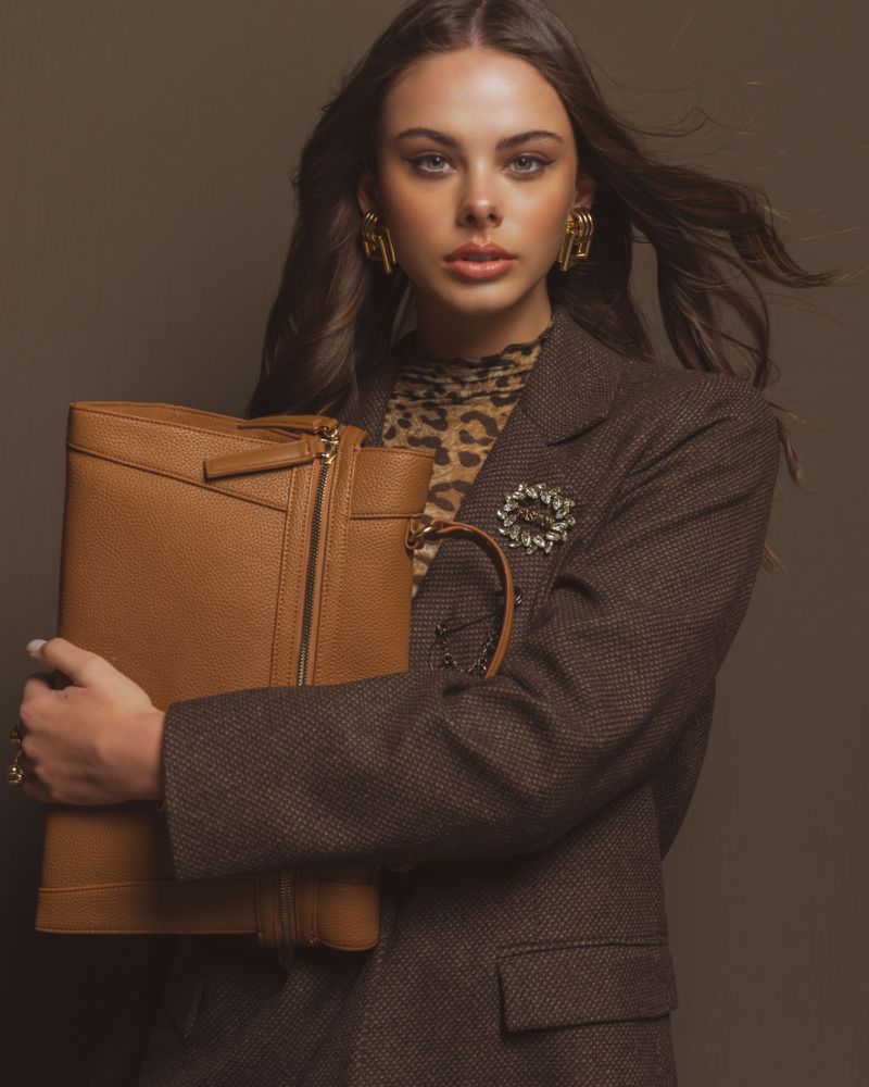 Woman in blazer holds leather handbag in studio. She wears gold earrings and a patterned top against a neutral backdrop.