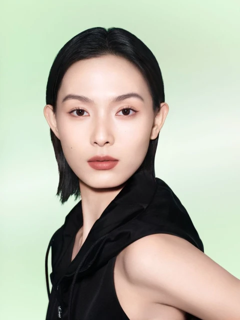 Close-up of a young Asian woman with dark hair and neutral makeup, wearing a black hooded vest against a light green gradient backdrop.