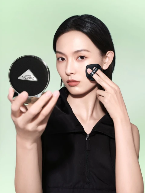 A woman applies makeup with a black applicator pad, holding a compact with the Prada logo, against a soft green background.