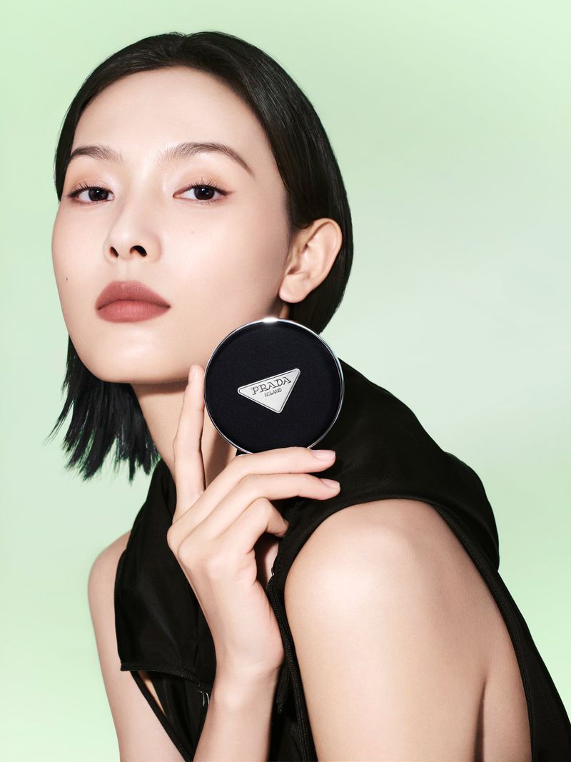 Asian woman showcases Prada compact, fair skin, short black hair, black top against pastel green background in a beauty advertisement.