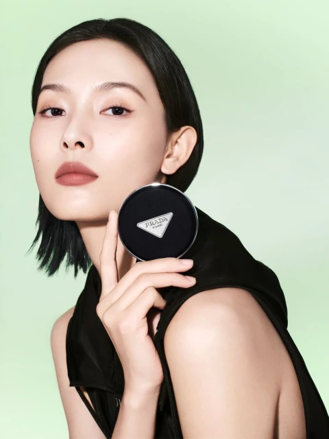 Asian woman showcases Prada compact, fair skin, short black hair, black top against pastel green background in a beauty advertisement.