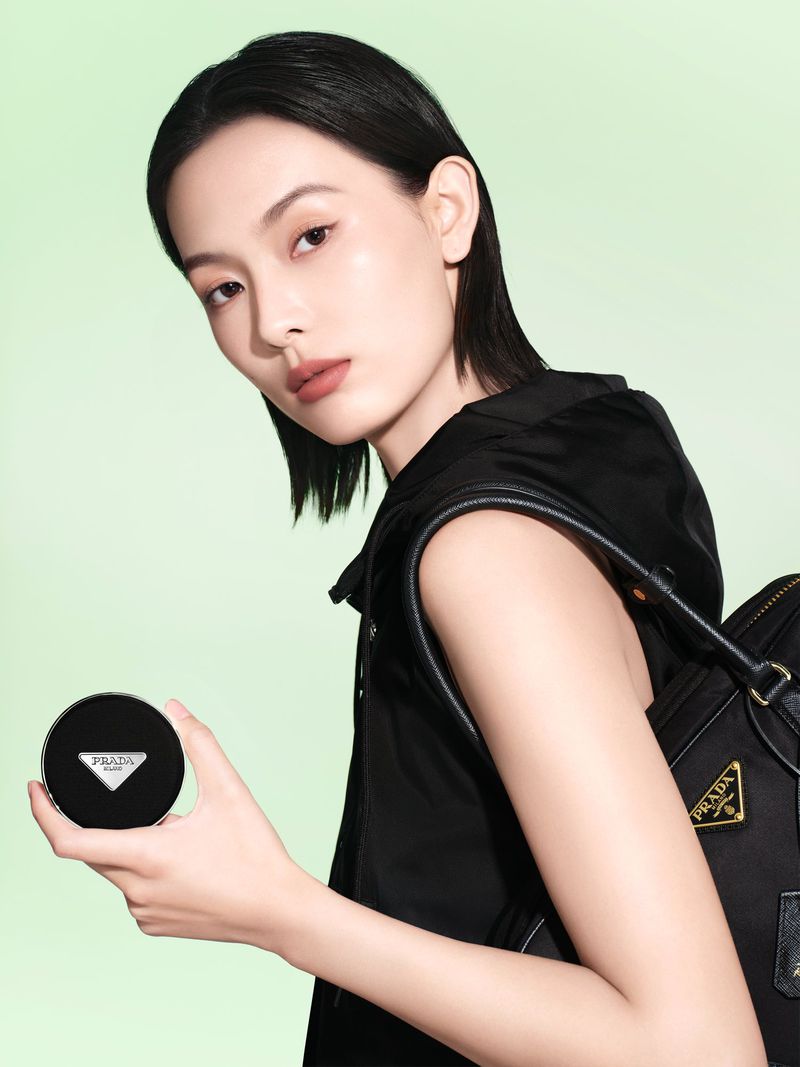 Beauty shot of a woman holding Prada foundation compact, wearing a black vest and a black shoulder bag against a lime green background.