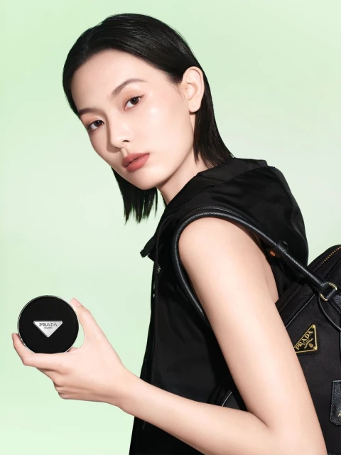 Beauty shot of a woman holding Prada foundation compact, wearing a black vest and a black shoulder bag against a lime green background.