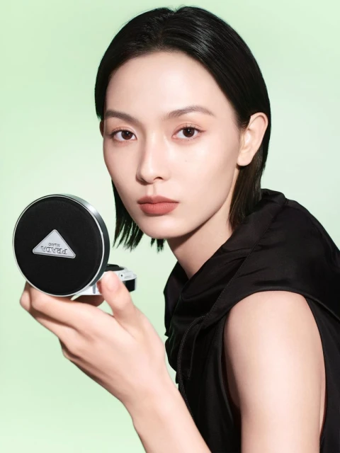 A woman with short black hair holds a black and silver Prada compact in a beauty advertisement against a light green background.