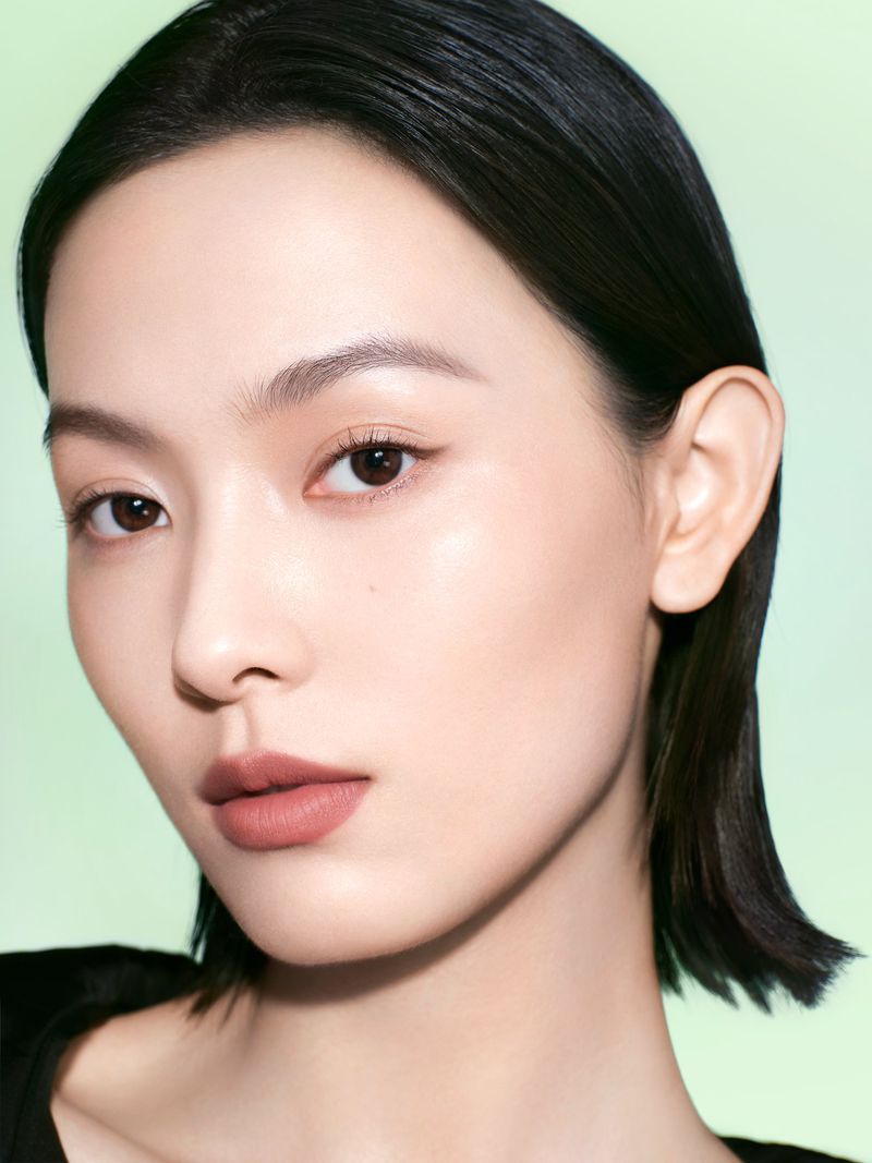 Portrait of an Asian woman with short black hair, light skin, and nude makeup on a pale green background in a beauty studio shot.