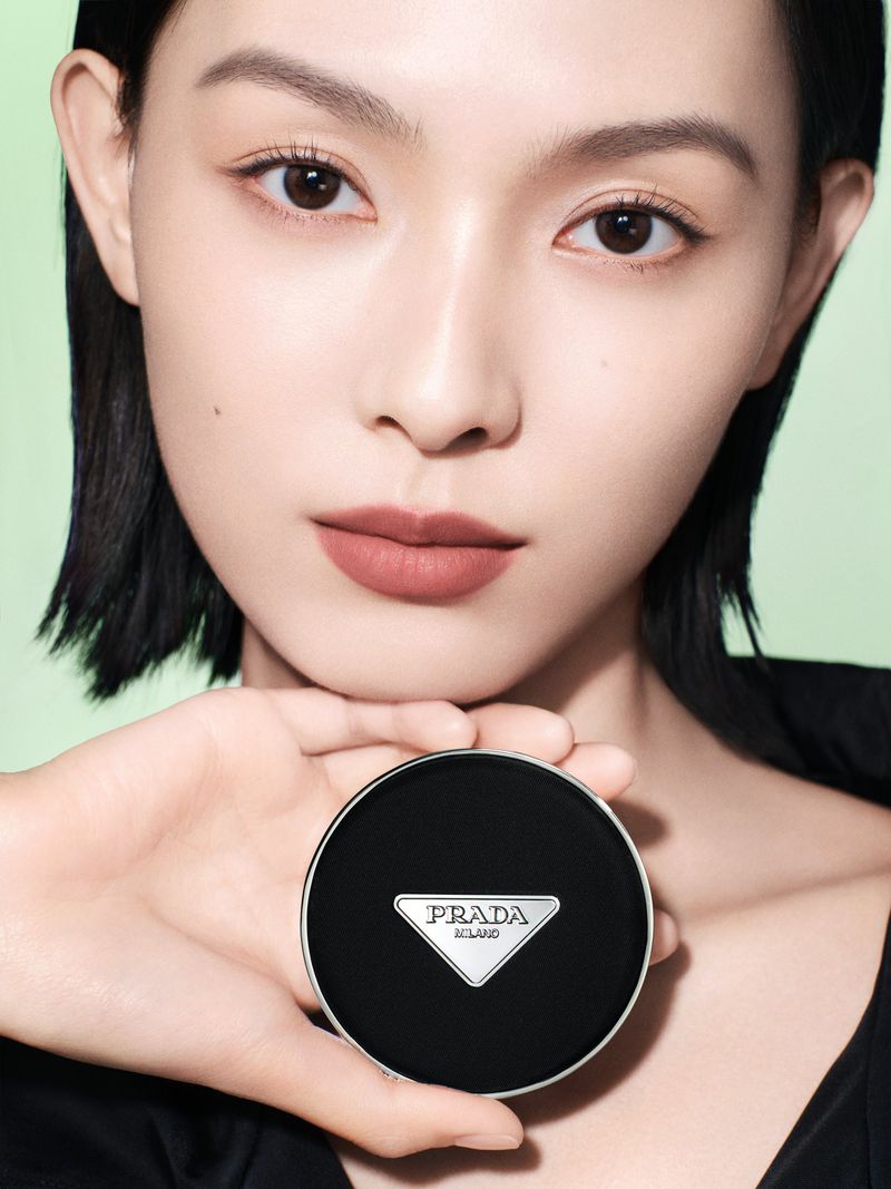 Asian woman holding a Prada compact case close to her face in a beauty shot. The background is a pale green colour.