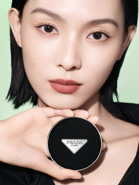 Asian woman holding a Prada compact case close to her face in a beauty shot. The background is a pale green colour.