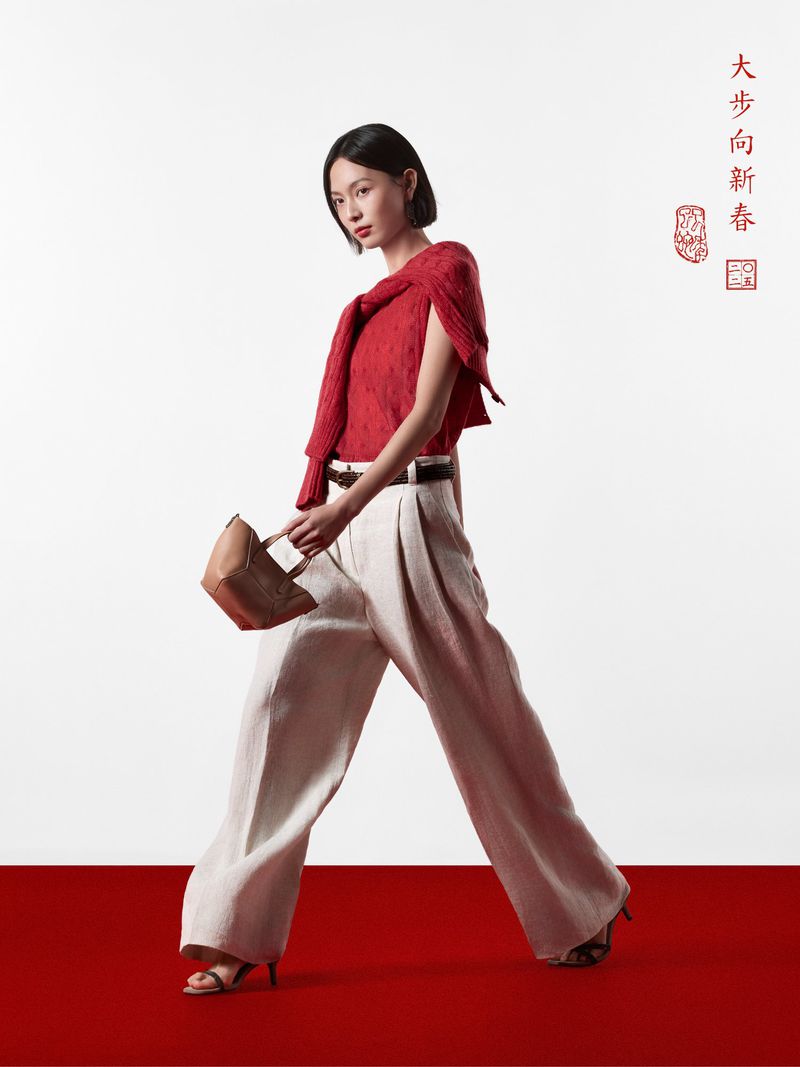 Fashionable woman walks in red top, tan pants, and heels, carrying handbag against a white backdrop with red floor and Chinese text.
