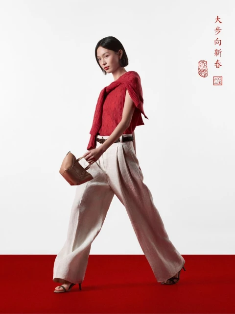 Fashionable woman walks in red top, tan pants, and heels, carrying handbag against a white backdrop with red floor and Chinese text.